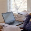 A teacher thinks at her laptop, surrounded by stacks of papers